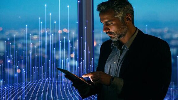 A employee looking at his tab standing near the glass window of the corporate office overlaid with graphics of transmission of nodes 