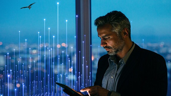 A professional working on his tab standing next to glass window in the modern corporate building overlaid with graphics of transmission nodes