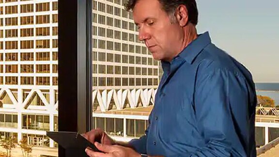 A person standing infront of the glass window of a tall building looking at a tab in his hand