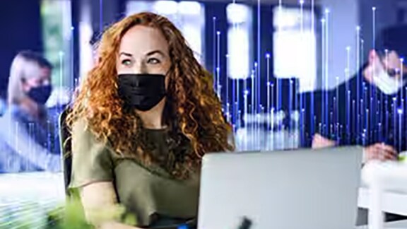 Lady employee with a mask sitting in office looking away from laptop overlaid with graphics of transmission nodes