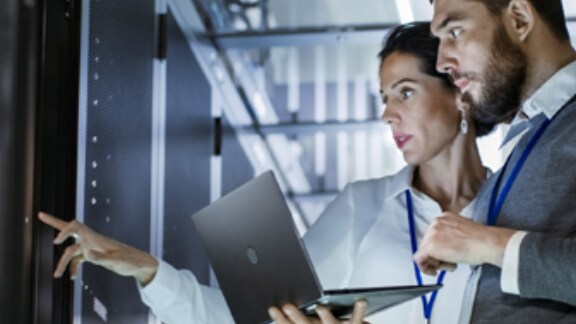 A man holding a laptop standing next to a woman pointing at a control panel