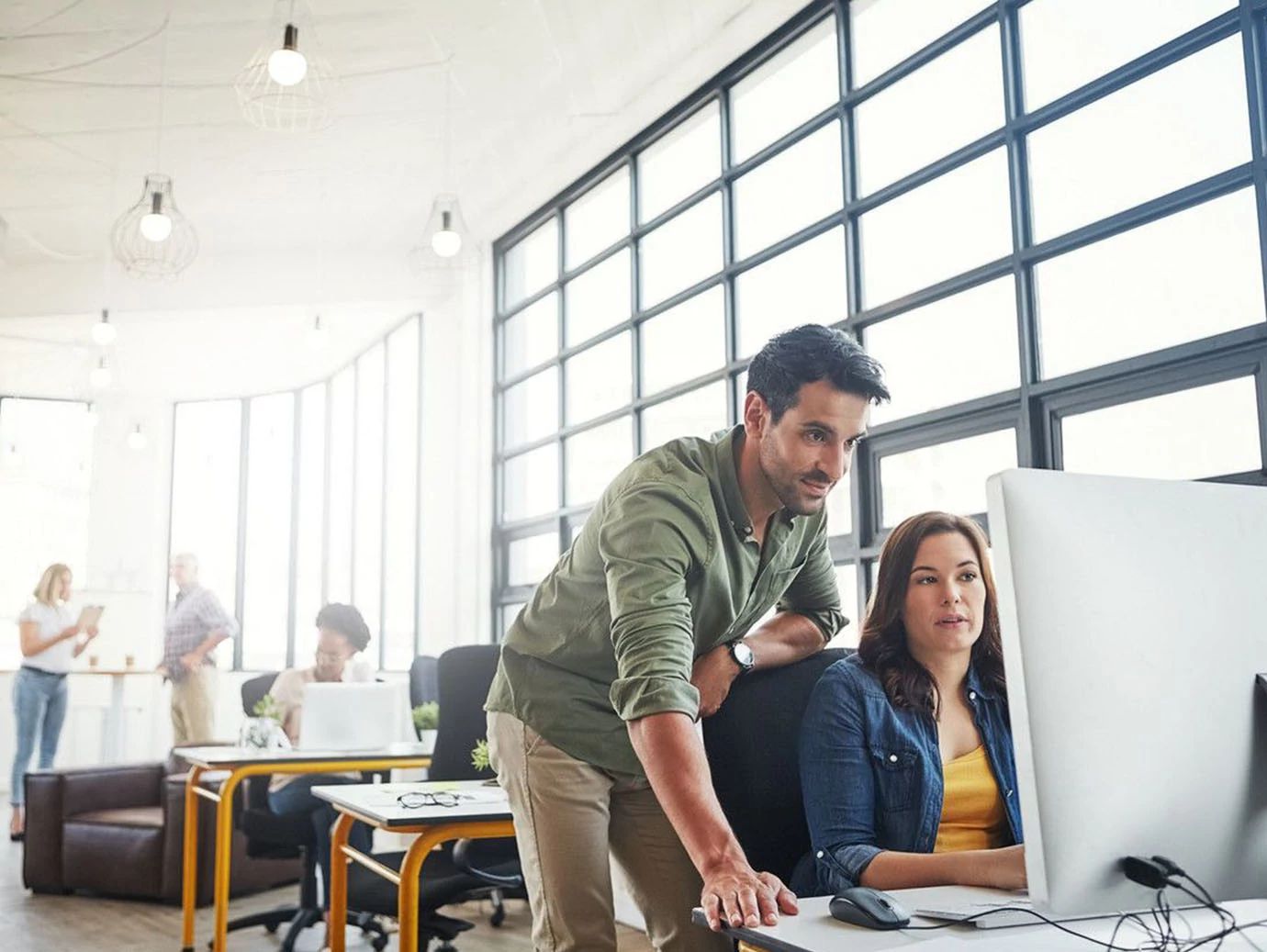 A man guiding a woman at a desktop, while other employees are seated at work behind them