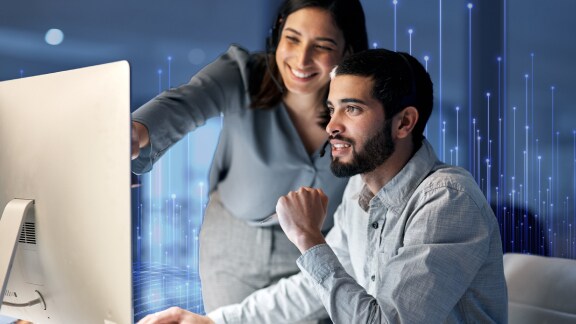 A man seated and looking at a monitor, while a woman stands behind him and points at the screen while smiling